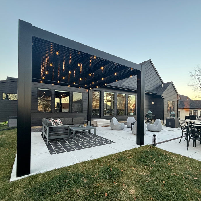 A somewhat faraway view of a black vinyl pergola with clean, squared-off corners covering a concrete patio and patio furniture in the back yard of a dark-colored home