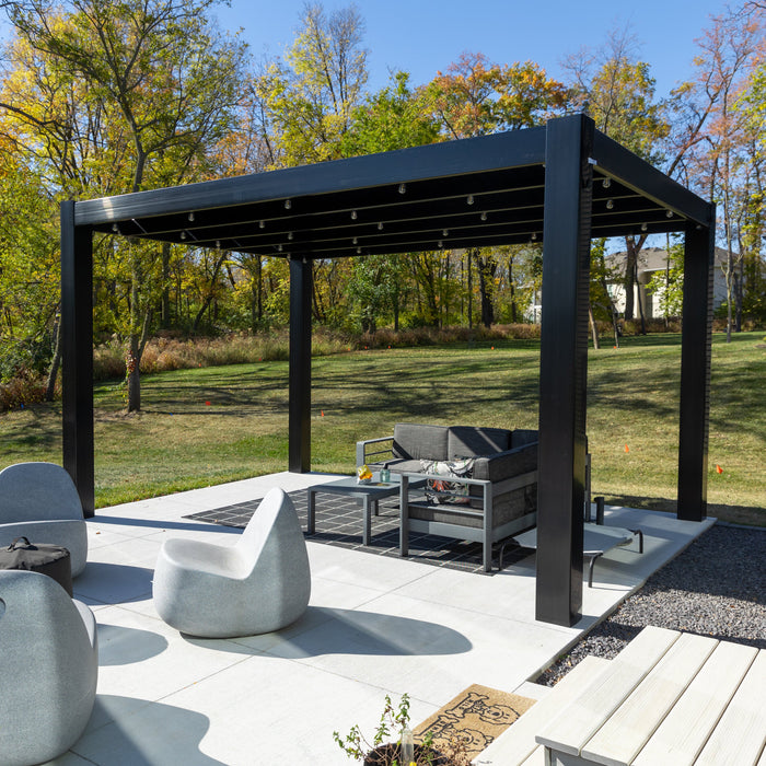 A black contemporary pergola at the edge of a patio, with stone seating around a firepit in the foreground and grass and trees in the background