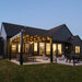 A distant view of the back of a home with a black vinyl pergola over a cement patio, with lights hanging from the roof of the pergola to provide light in the evening