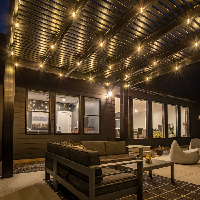 A night view of a black modern free-standing pergola covering a cement patio behind a house, with large lights hanging from the rafters of the pergola to provide a nice ambiance in the dark