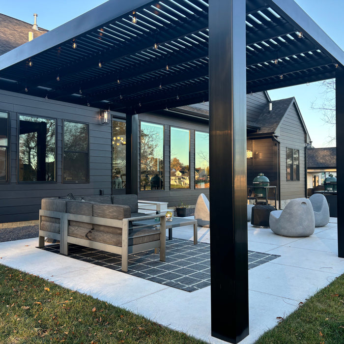 Picture of a black modern-style pergola with clean, squared-off corners against the back of a dark-colored house, with patio furniture underneath the pergola