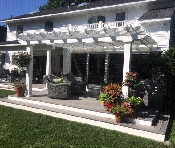 Picture of a white traditional pergola attached to the back of a white home covering a slightly raised Trex deck with patio furniture and plants