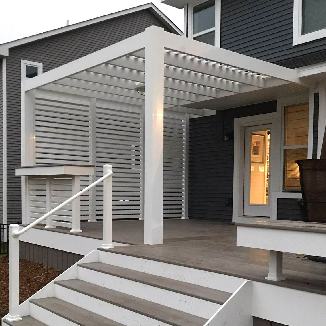 Picture of a 2-post white vinyl wall-mounted contemporary pergola covering the back deck of a dark-colored house