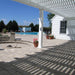 Picture taken underneath a white traditional pergola attached to a white house near a nice circular firepit and a swimming pool in the background