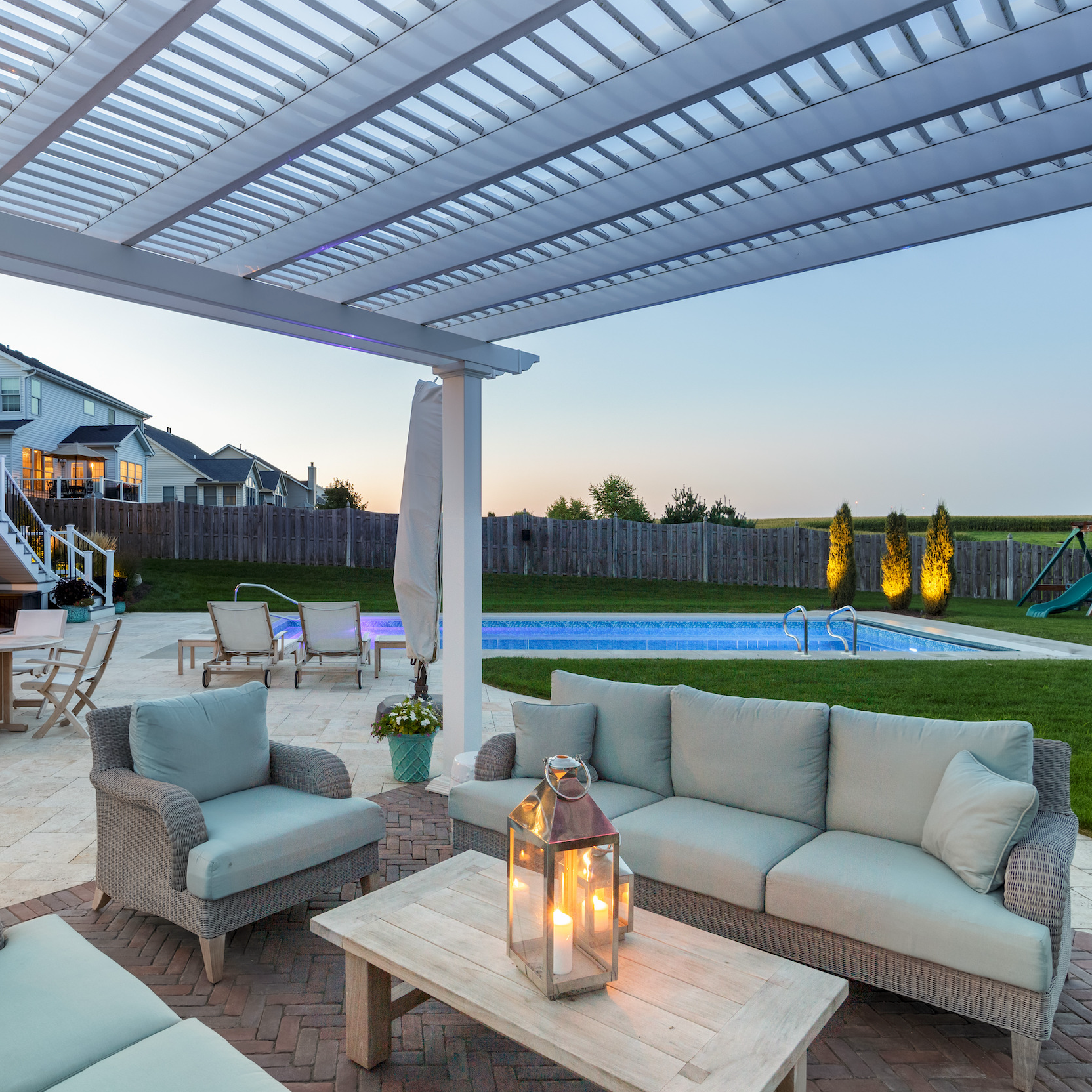 View from underneath a white traditional pergola with 3 outdoor patio couches and a swimming pool and home in the background