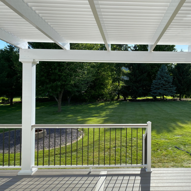 Picture taken from underneath a white vinyl pergola covering a trex deck with grass and trees in the background