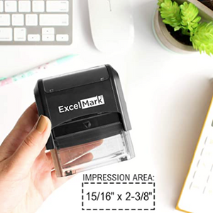 Picture of a hand holding an ExcelMark branded self-inking stamp with various office and desk supplies in the background