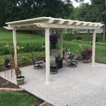 Sandstone tan 4-post traditional pergola with overhangs at the edge of a cement patio surrounded by flowers and potted plants with trees and a home in the background