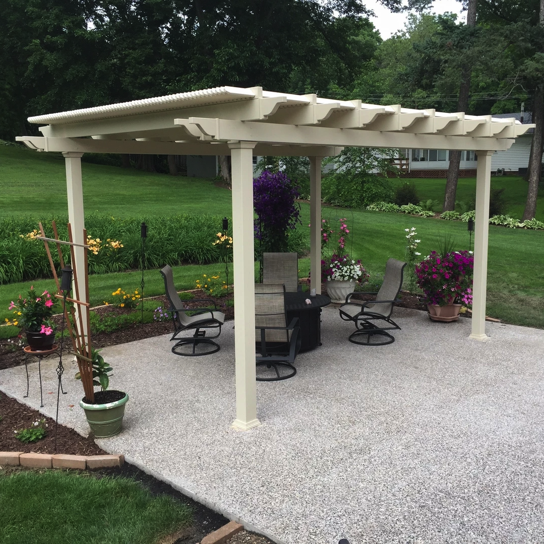 Sandstone tan 4-post traditional pergola with overhangs at the edge of a cement patio surrounded by flowers and potted plants with trees and a home in the background